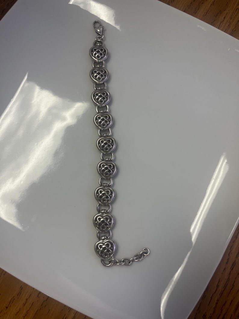 Puede incluir: Una pulsera plateada con eslabones en forma de coraz&oacute;n. Cada coraz&oacute;n presenta un dise&ntilde;o calado intrincado. La pulsera se muestra sobre una superficie blanca, resaltando sus delicados detalles y el cierre de cadena.