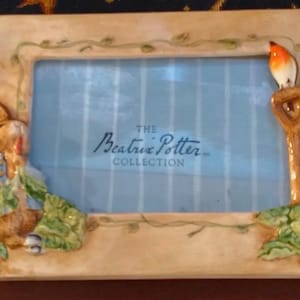 May include: A rectangular ceramic picture frame from The Beatrix Potter Collection featuring a sculpted Peter Rabbit on the left and a small orange-breasted bird perched on a garden tool on the right, with painted green vines on the frame border.