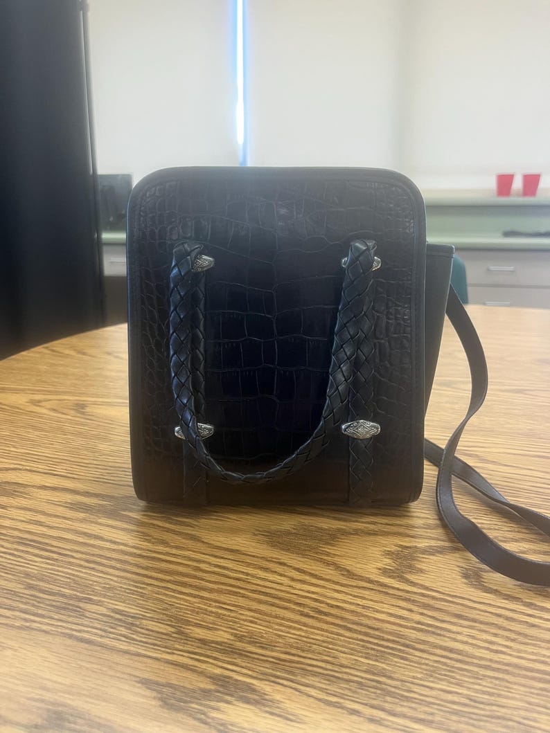 May include: Black square handbag with a crocodile texture. The bag has braided handles and silver-toned hardware. A long, thin strap is attached for shoulder or crossbody wear. The bag sits on a wooden table.