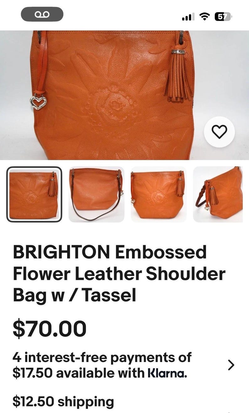 May include: An orange shoulder bag featuring an embossed floral pattern on the front, a matching leather tassel attached to the side, and a silver-toned heart-shaped charm hanging from a strap. The bag has a structured shape and a smooth finish.