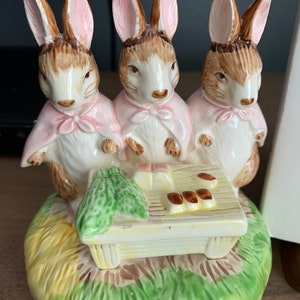 May include: A ceramic musical figurine of three bunnies wearing pink cloaks and sitting on a green and yellow base. The bunnies are playing a small white piano with brown keys.