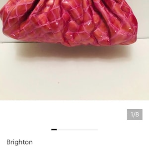 May include: A pink coin purse clutch with a textured, crocodile-like pattern. The purse has a pink frame and a silver chain handle. The clasp features a decorative ball with a pink ribbon design. The purse is from the brand Brighton.