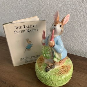 May include: A ceramic figurine of Peter Rabbit holding carrots and a bundle of greens, standing on a grassy base. Beside it is a copy of "The Tale of Peter Rabbit" book by Beatrix Potter.