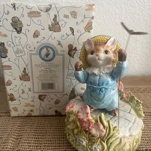 May include: A ceramic Tom Kitten musical figurine from The World of Beatrix Potter, wearing a blue outfit and yellow straw hat, standing on a floral base with a butterfly on a wire. The item is accompanied by its original patterned cardboard box.