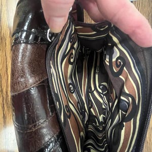 May include: A brown leather handbag with a glossy finish. The bag features a patchwork design with varying shades of brown and a patterned interior lining. The zipper is open, revealing the inside of the bag.