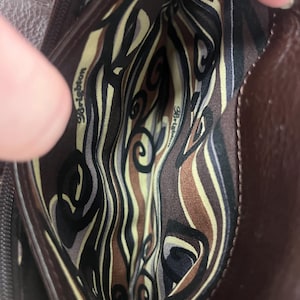 May include: A brown leather handbag interior with a patterned fabric lining. The lining features a swirl design in black, tan, and olive green. The brand name "Brighton" is visible on the lining.