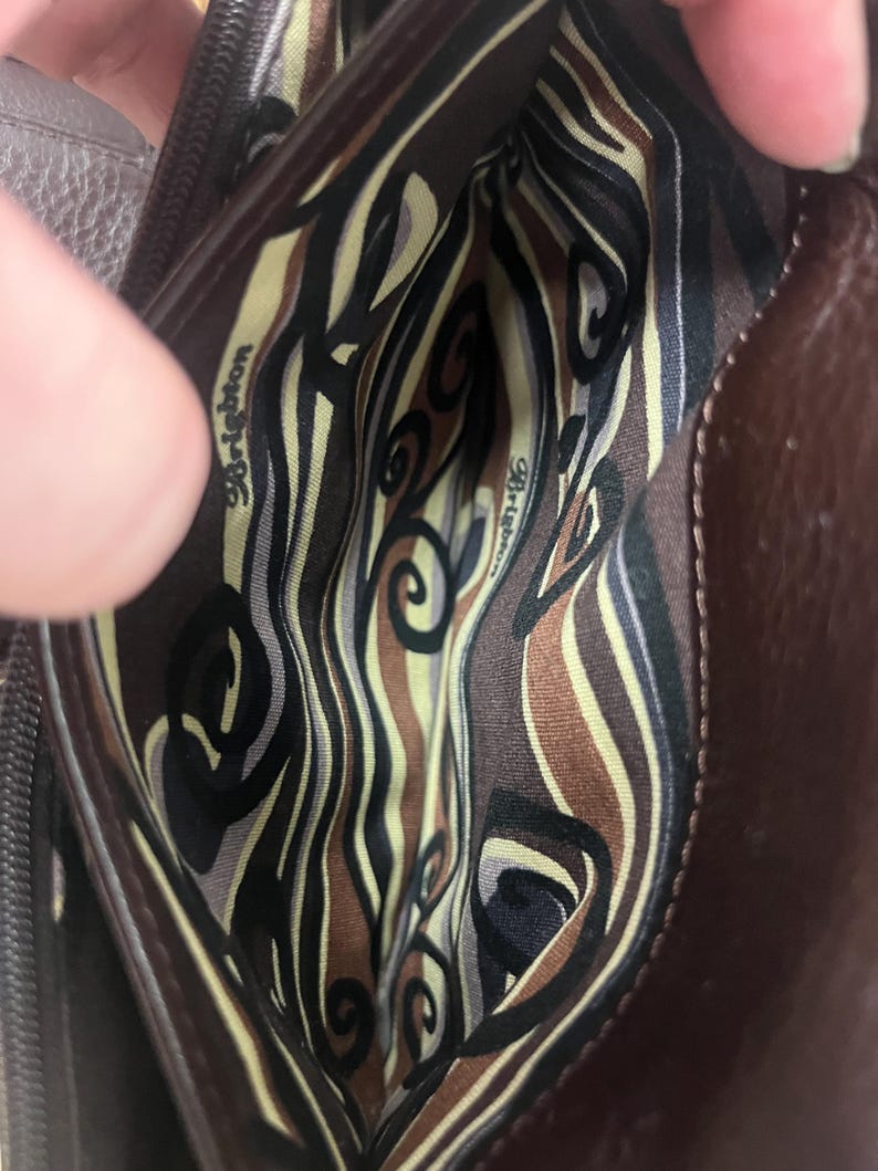 May include: A brown leather handbag interior with a patterned fabric lining. The lining features a swirl design in black, tan, and olive green. The brand name "Brighton" is visible on the lining.