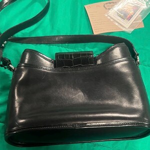 May include: Black leather shoulder bag with a crocodile-textured flap closure. The bag has a long strap and a rounded bottom. The bag is on a green surface.