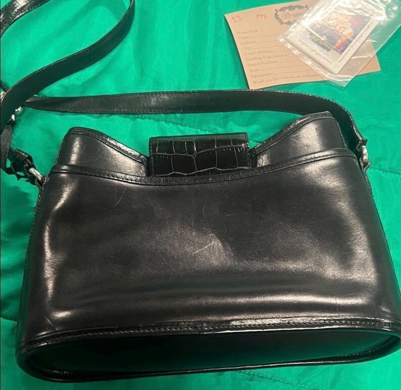 May include: Black leather shoulder bag with a crocodile-textured flap closure. The bag has a long strap and a rounded bottom. The bag is on a green surface.