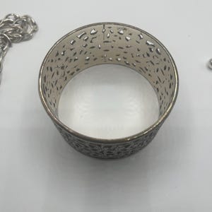 May include: Silver-toned metal cuff bracelet with an openwork design. The bracelet has a cylindrical shape with intricate cut-out patterns. A silver chain is partially visible in the background.