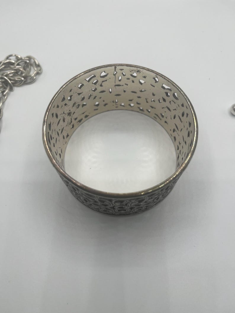 May include: Silver-toned metal cuff bracelet with an openwork design. The bracelet has a cylindrical shape with intricate cut-out patterns. A silver chain is partially visible in the background.