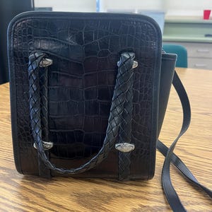 May include: Black square handbag with a textured crocodile pattern. The bag features braided handles and silver-toned hardware. A long, thin strap is attached for shoulder or crossbody wear.