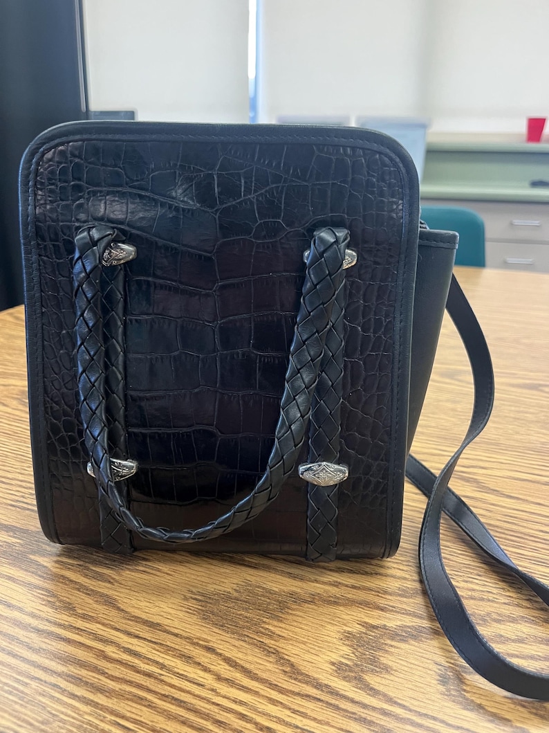 May include: Black square handbag with a textured crocodile pattern. The bag features braided handles and silver-toned hardware. A long, thin strap is attached for shoulder or crossbody wear.