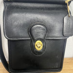 May include: A black leather shoulder bag featuring a rectangular flap, a brass turn-lock closure, and a matching leather hang tag attached by a gold-toned chain. The bag includes a long adjustable shoulder strap and gold-toned hardware accents.