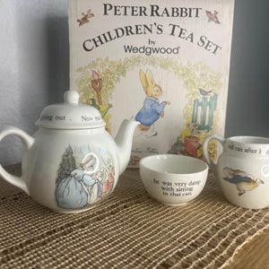 May include: A Peter Rabbit Children's Tea Set by Wedgwood. The set includes a white teapot, a small bowl, and a creamer. The teapot has a lid and a handle. The bowl and creamer have text. The box behind the set has a Peter Rabbit illustration.