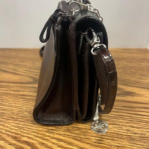 May include: A dark brown leather crossbody bag with a silver chain strap and a decorative charm. The bag has a flap closure and a textured finish. The bag is sitting on a wooden surface.