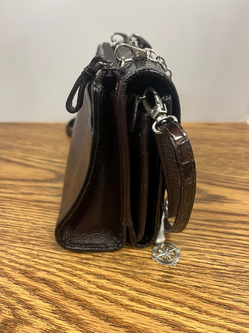 May include: A dark brown leather crossbody bag with a silver chain strap and a decorative charm. The bag has a flap closure and a textured finish. The bag is sitting on a wooden surface.