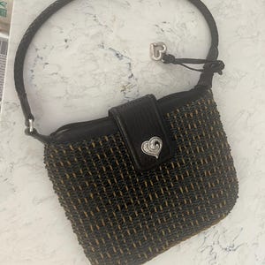 Puede incluir: Un bolso tejido negro y marr&oacute;n con un asa trenzada. El bolso presenta una solapa de cuero negro con un cierre plateado en forma de coraz&oacute;n. Un dije de coraz&oacute;n plateado cuelga del asa.