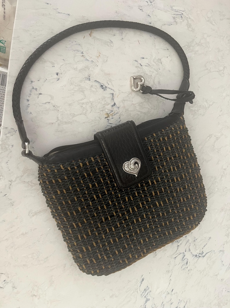 Puede incluir: Un bolso tejido negro y marr&oacute;n con un asa trenzada. El bolso presenta una solapa de cuero negro con un cierre plateado en forma de coraz&oacute;n. Un dije de coraz&oacute;n plateado cuelga del asa.