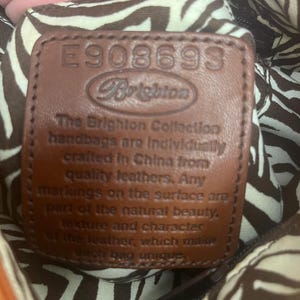 May include: A brown leather patch with the embossed text E908693 and Brighton, followed by a statement about the brand's craftsmanship. The patch is set against a fabric lining featuring a brown and white zebra stripe pattern.