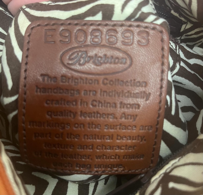 May include: A brown leather patch with the embossed text E908693 and Brighton, followed by a statement about the brand's craftsmanship. The patch is set against a fabric lining featuring a brown and white zebra stripe pattern.