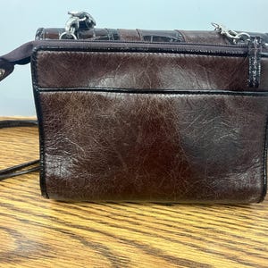 May include: A small, brown leather crossbody bag with a textured finish. The bag features a top zip, a black trim, and a silver chain strap. The bag is sitting on a wooden surface.