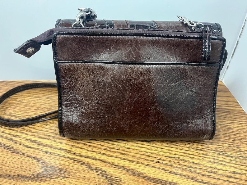 May include: A small, brown leather crossbody bag with a textured finish. The bag features a top zip, a black trim, and a silver chain strap. The bag is sitting on a wooden surface.