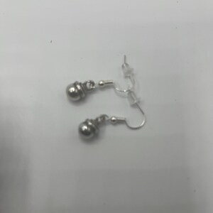 May include: A pair of silver-colored earrings. Each earring features a small, round, textured bead suspended from a curved ear wire. The earrings are displayed against a plain, light-colored background.