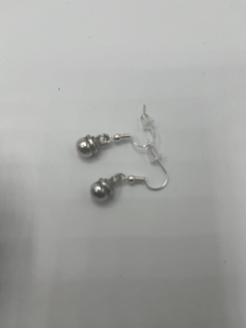 May include: A pair of silver-colored earrings. Each earring features a small, round, textured bead suspended from a curved ear wire. The earrings are displayed against a plain, light-colored background.