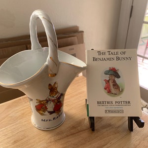 May include: A white porcelain basket with a gold handle and a depiction of Mrs. Rabbit and her children from Beatrix Potter's The Tale of Peter Rabbit. The basket is next to a copy of The Tale of Benjamin Bunny by Beatrix Potter.