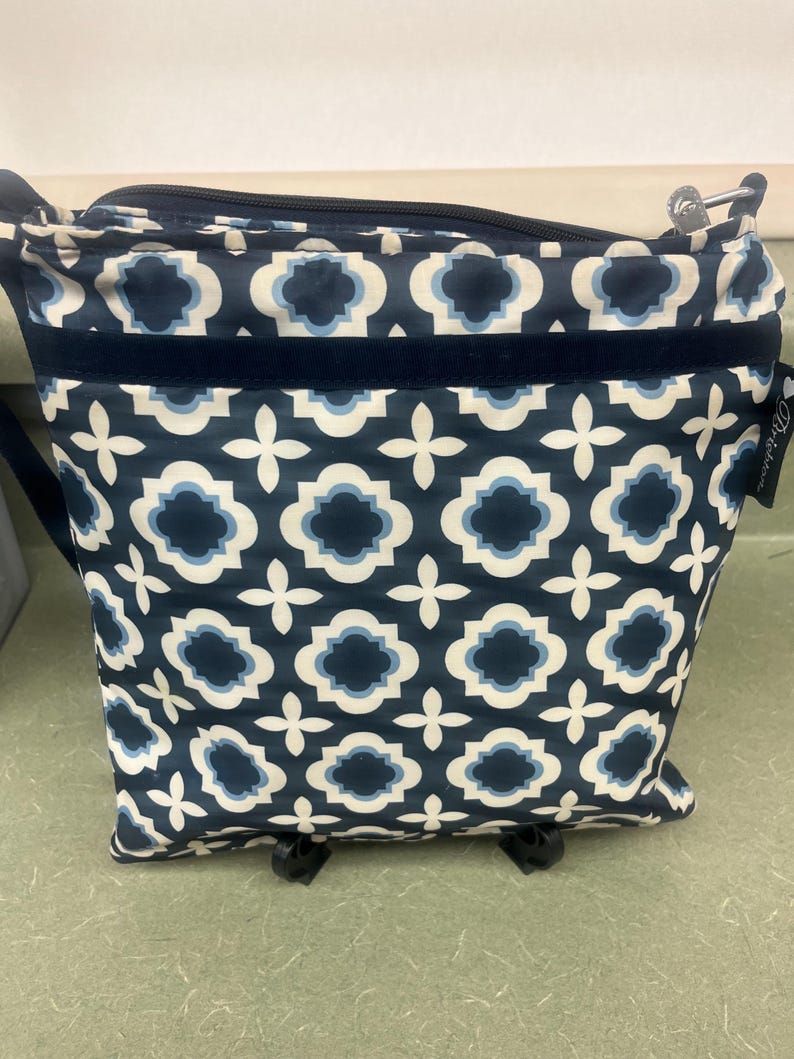 May include: A navy blue crossbody bag with a geometric pattern in white and light blue. The bag has a zippered top, a navy blue velvet stripe, and a silver-toned metal zipper pull. The bag has a black strap and a small tag that says "Brighton".
