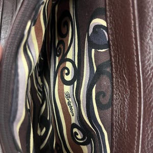 May include: An open brown leather handbag, revealing the patterned interior lining. The lining displays swirls and stripes in brown, black, and cream. The brand name "Brighton" is printed on the lining.