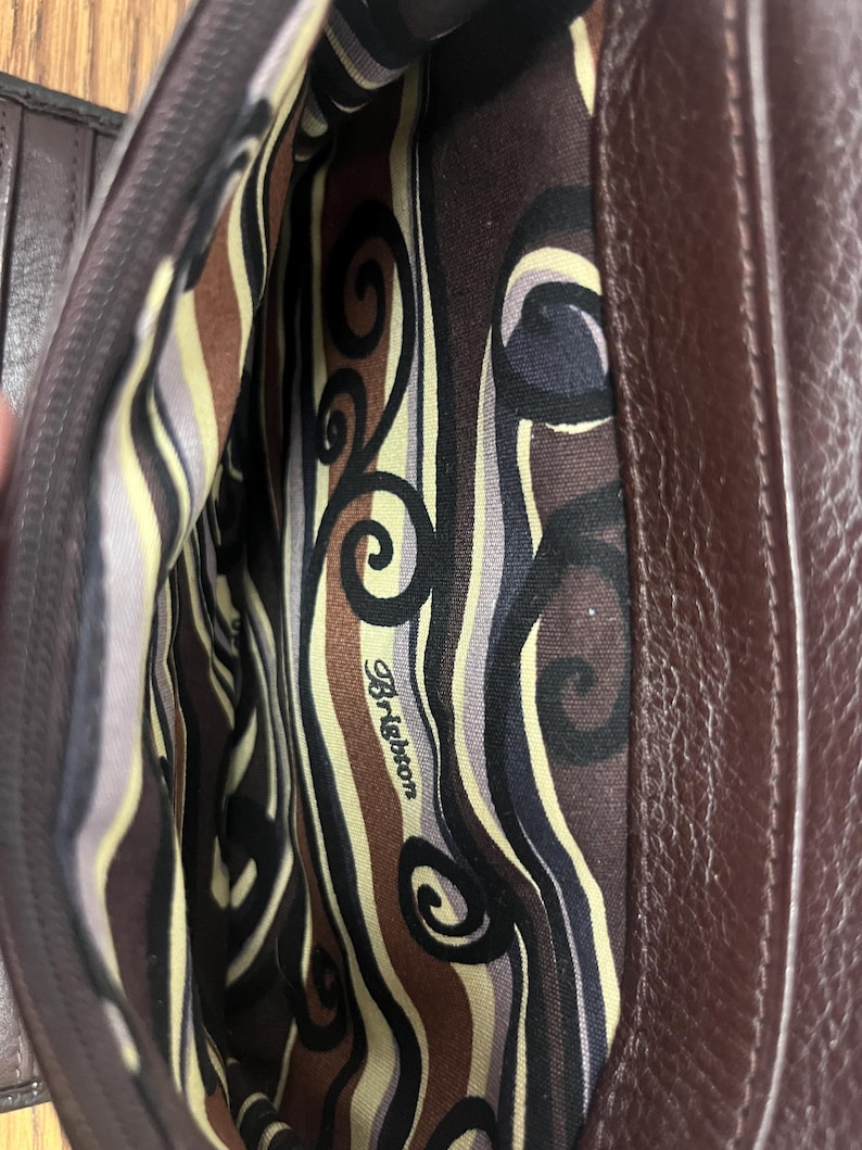 May include: An open brown leather handbag, revealing the patterned interior lining. The lining displays swirls and stripes in brown, black, and cream. The brand name "Brighton" is printed on the lining.