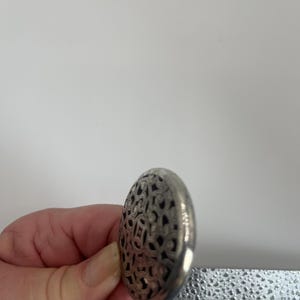 May include: A silver-colored, oval-shaped object with an intricate openwork design. The object is held in a hand, and the design appears to be a repeating pattern of swirls and shapes. The object is likely a decorative element or a piece of jewelry.