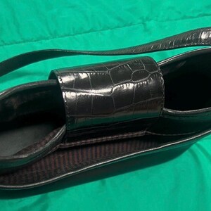 May include: Black handbag with a textured, crocodile-like pattern. The bag has a long strap and a rectangular shape with a central handle. The interior lining is a dark brown with vertical stripes.