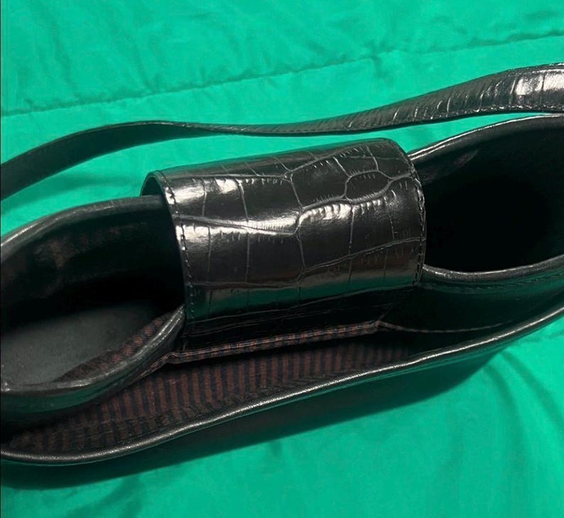 May include: Black handbag with a textured, crocodile-like pattern. The bag has a long strap and a rectangular shape with a central handle. The interior lining is a dark brown with vertical stripes.