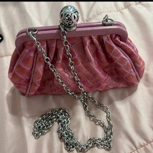 May include: A pink clutch purse with a silver chain strap. The purse has a textured, crocodile-like pattern and a decorative silver ball clasp. The frame is a lighter shade of pink.