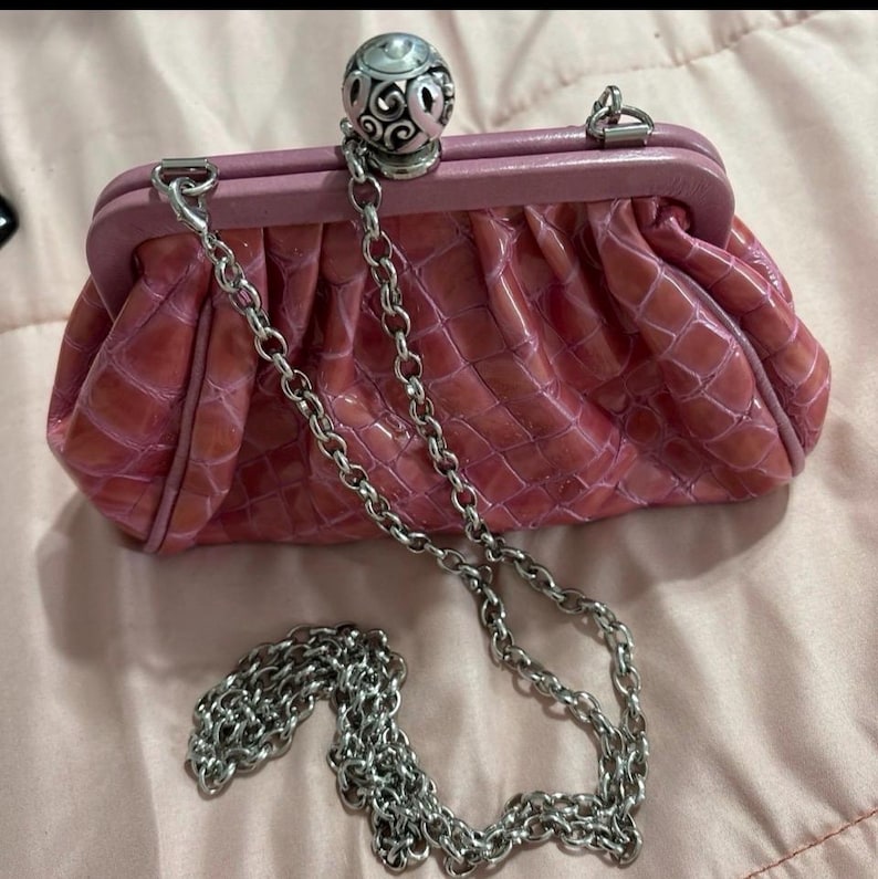 May include: A pink clutch purse with a silver chain strap. The purse has a textured, crocodile-like pattern and a decorative silver ball clasp. The frame is a lighter shade of pink.