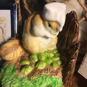 May include: A ceramic figurine of a squirrel wearing a white chef's hat, perched on a tree stump with green foliage. The squirrel has tan and cream-colored fur. The base is brown with green grass and leaves.
