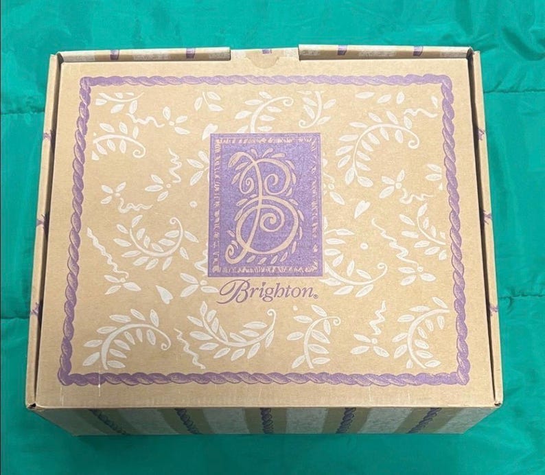 May include: A rectangular cardboard box with a beige background and a purple border. The box features a central purple square with a stylized letter "B" and the word "Brighton" in a script font. The box is decorated with white floral and vine patterns.