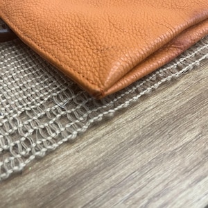 May include: A close-up view of a folded orange leather item with a visible pebbled texture. The material features clean, straight stitching along the edge, showcasing the natural grain and the vibrant, warm orange hue of the leather surface.