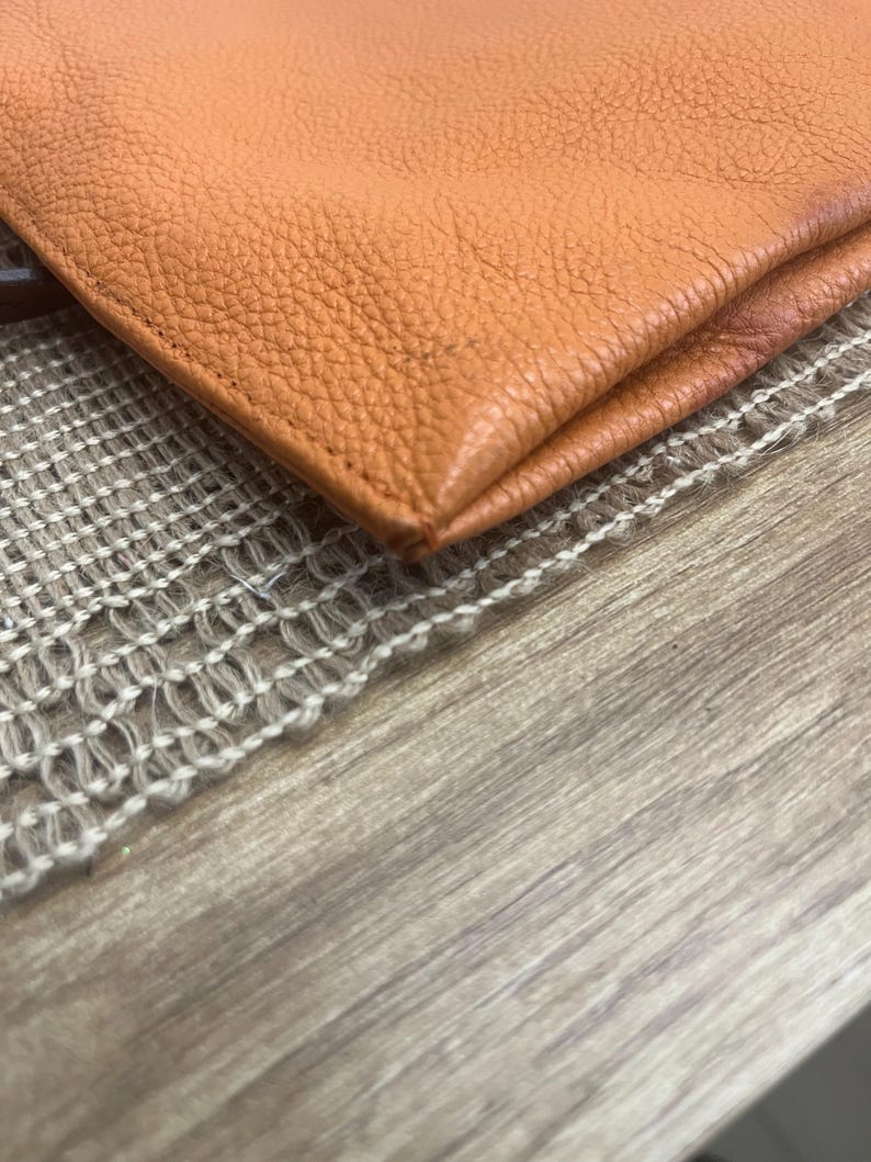 May include: A close-up view of a folded orange leather item with a visible pebbled texture. The material features clean, straight stitching along the edge, showcasing the natural grain and the vibrant, warm orange hue of the leather surface.