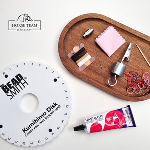 Op de afbeelding: Een witte Kumihimo-schijf met zwarte nummers en de tekst "the BEAD SMITH" en "Kumihimo Disk Create your own braided cord!" Een houten dienblad met een roze doek, een spoel garen, een naald, een metalen sluiting, een metalen gewicht met een haak, een tandenstoker en een klein zakje kleurrijke elastiekjes. Een tube Hasulith-lijm.