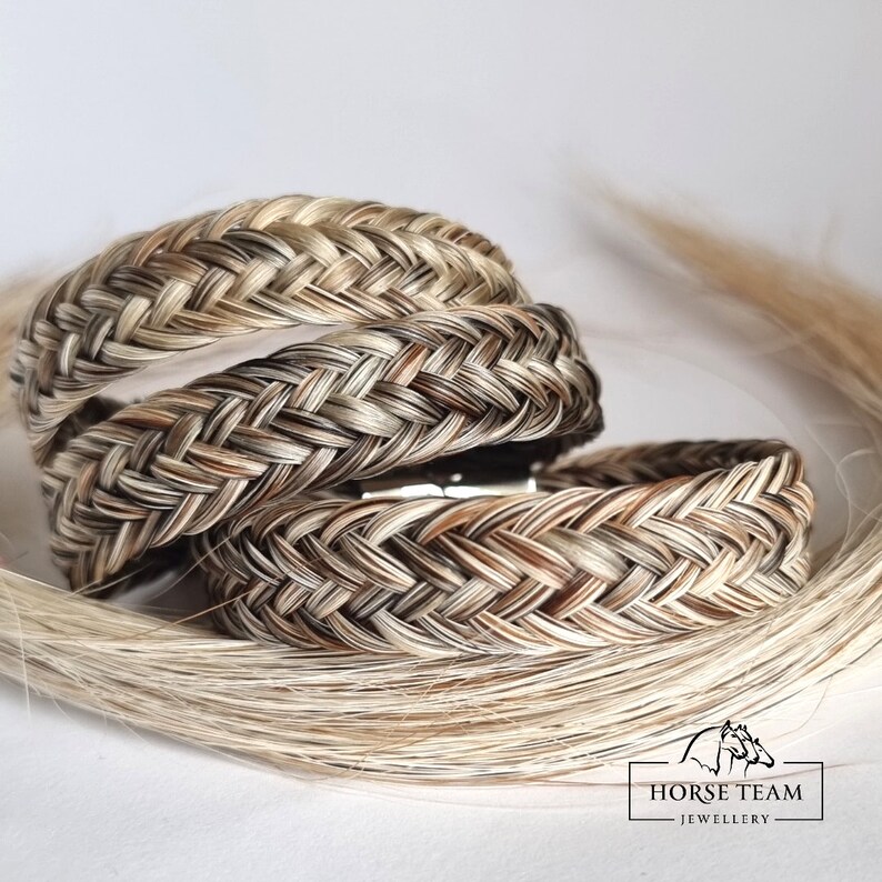 May include: Three braided horsehair bracelets in shades of brown, white, and black. The bracelets are stacked on top of each other and are made with a traditional braid pattern. The image also includes a strand of white horsehair.