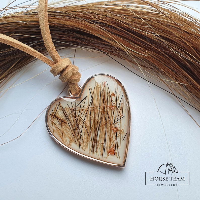 May include: A heart-shaped pendant necklace with a brown leather cord. The pendant is filled with brown horse hair and has a silver-toned metal frame. The pendant is hanging against a background of brown horse hair.