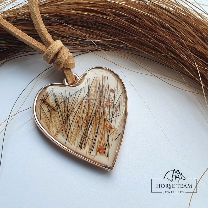 May include: A heart-shaped pendant necklace with a brown leather cord. The pendant is filled with horse hair and has a gold-colored metal frame. The pendant is surrounded by a pile of brown horse hair.
