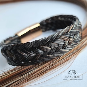 May include: A black, brown, and white braided horsehair bracelet with a gold clasp. The bracelet is made by Horse Team Jewellery.