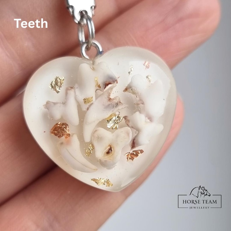 May include: A white heart-shaped pendant with a silver chain. The pendant contains several small white teeth and gold and copper flakes. The pendant is labelled "Teeth".