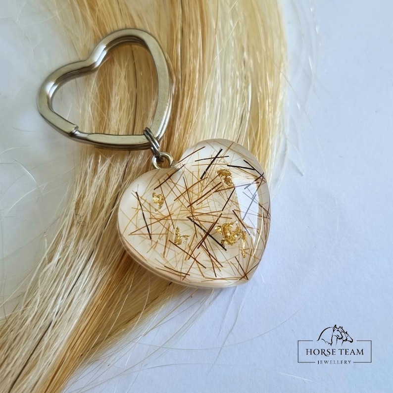May include: A heart-shaped keyring with a clear resin design. The resin contains strands of blonde hair and gold flakes. The keyring is attached to a silver metal ring.