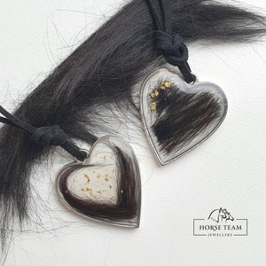 May include: Two silver heart-shaped pendants with black horse hair encased in clear resin. Each pendant has gold flakes embedded in the resin. The pendants are attached to black cords.